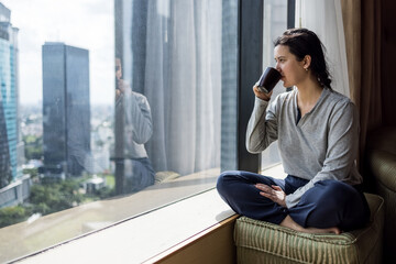 Young woman holding coffee cup, wearing pajama and looking at cityscape through the window in luxury penthouse apartment or hotel room - obrazy, fototapety, plakaty
