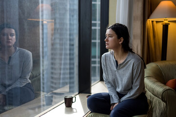 Young woman holding coffee cup, wearing pajama and looking at cityscape through the window in luxury penthouse apartment or hotel room - 619149296