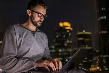 Handsome man using laptop in penthouse or luxury hotel room. Businessman in skyscraper apartment against night city background working late at nigh.
