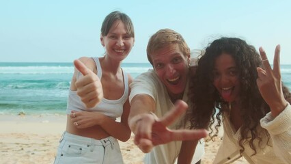 Young cheerful positive friends in summer clothes shout joyfully recording video for blog about travel or life on tropical islands and invite you to come for vacation stands on sea beach.