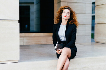 Confidently posed female model with wavy hair, red lips, formal attire, holding tablet, crossing...
