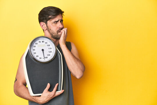 Athletic man with scale, on a yellow studio backdrop biting fingernails, nervous and very anxious.