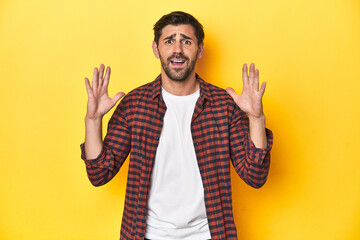 Caucasian man in red checkered shirt, yellow backdrop screaming to the sky, looking up, frustrated.