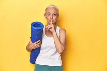 Fitness woman with yoga mat, sportswear on yellow backdrop keeping a secret or asking for silence.