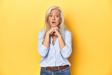 Middle-aged Caucasian woman in blue shirt, yellow studio praying for luck, amazed and opening mouth...