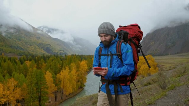 Caucasian backpacker with rucksack use smartphone online, hold device, touch, scroll and tap. Young male person in equipment at extreme vacation walk and use cellphone. Beautiful autumn natural area
