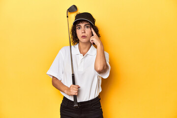 Athletic Caucasian woman with curly hair golfing in studio pointing temple with finger, thinking, focused on a task.