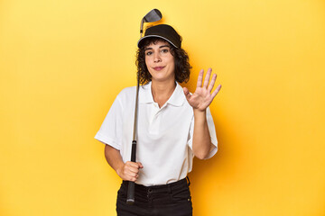 Athletic Caucasian woman with curly hair golfing in studio smiling cheerful showing number five...