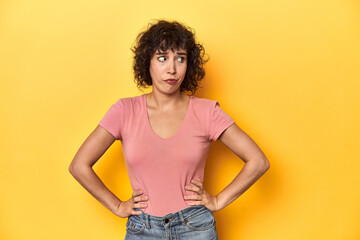 Curly-haired Caucasian woman in pink t-shirt confused, feels doubtful and unsure.