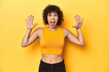 Curly-haired Caucasian woman in yellow top receiving a pleasant surprise, excited and raising hands.