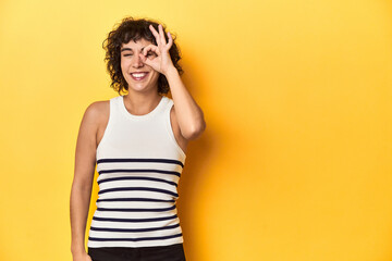 Caucasian curly-haired woman in white tank-top excited keeping ok gesture on eye.