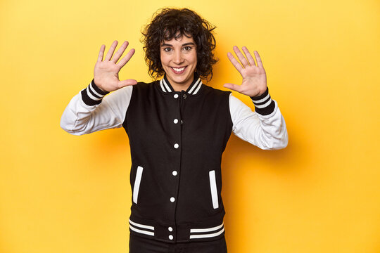 Curly-haired Caucasian woman in baseball jacket showing number ten with hands.