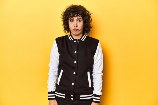 Curly-haired Caucasian woman in baseball jacket blows cheeks, has tired expression. Facial expression concept.