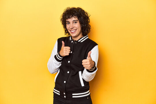 Curly-haired Caucasian woman in baseball jacket raising both thumbs up, smiling and confident.