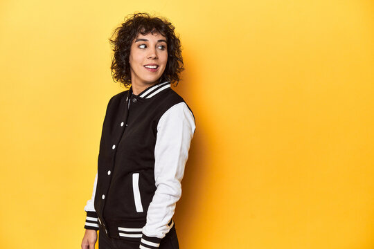 Curly-haired Caucasian woman in baseball jacket looks aside smiling, cheerful and pleasant.