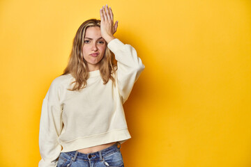 Young blonde Caucasian woman in a white sweatshirt on a yellow studio background, forgetting something, slapping forehead with palm and closing eyes.