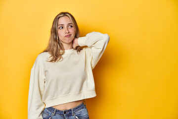 Young blonde Caucasian woman in a white sweatshirt on a yellow studio background, touching back of...