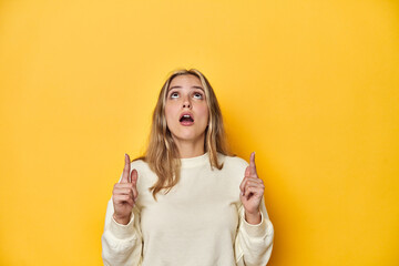 Young blonde Caucasian woman in a white sweatshirt on a yellow studio background, pointing upside...