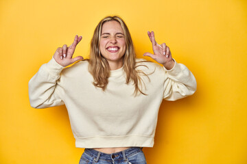 Young blonde Caucasian woman in a white sweatshirt on a yellow studio background, crossing fingers...