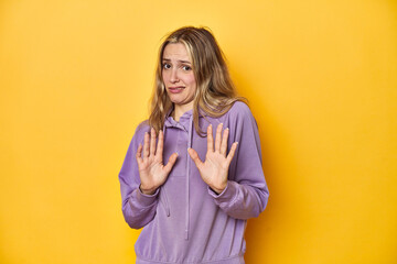 Young blonde Caucasian woman in a violet sweatshirt on a yellow background, rejecting someone...