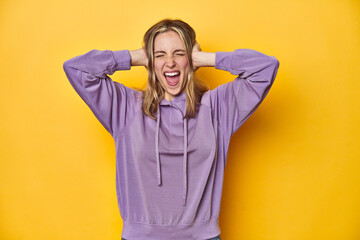 Young blonde Caucasian woman in a violet sweatshirt on a yellow background, covering ears with hands trying not to hear too loud sound.