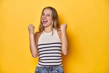 Young blonde Caucasian woman in a white tank top on a yellow studio background, cheering carefree...
