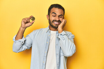 Young Latino man holding a Bitcoin coin, yellow studio background, covering ears with hands.