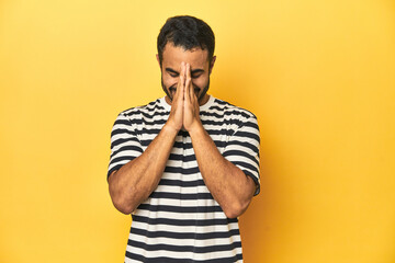 Casual young Latino man against a vibrant yellow studio background, holding hands in pray near...