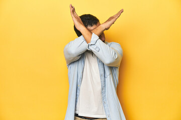 Casual young Latino man against a vibrant yellow studio background, keeping two arms crossed, denial concept.