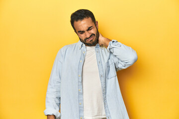 Casual young Latino man against a vibrant yellow studio background, having a neck pain due to stress, massaging and touching it with hand.