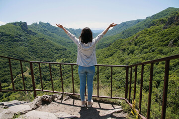 A young girl with a backpack enjoys the beauty of nature, looking at the view with outstretched arms at the top of the mountain