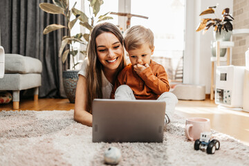 Happy middle age woman playing with her little son at home. They are using smart phone and laptop computer for playing, learning and online surfing. - obrazy, fototapety, plakaty