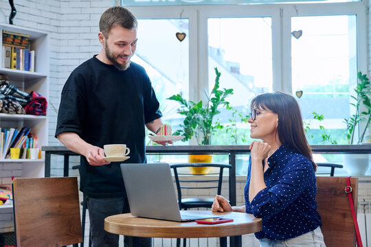 Young male waiter, owner of coffee shop serving cake and cup of coffee to female client