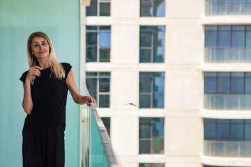 Portrait of young cute woman in black on skyscraper balcony with view of Dubai UAE, happy looking at camera. Smiling lady posing on terrace of tower block. Leisure activity concept. Copy ad text space