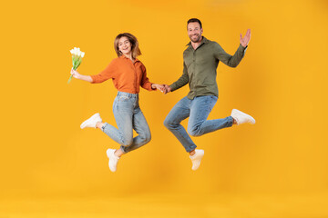 Fototapeta na wymiar Full length shot of happy european couple jumping high, woman holding fresh bunch flowers, man and woman in good mood isolated on yellow background