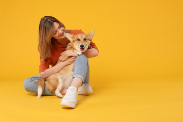 Fototapeta na wymiar Happy young caucasian woman holding a dog corgi, sitting on yellow studio background and embracing her pet, copy space, banner