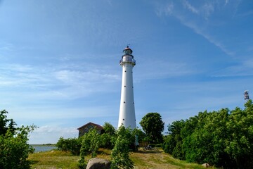 Kihnu island lighthouse in Estonia. Stand alone single white lighthouse stones green forest summer blue sky. - obrazy, fototapety, plakaty