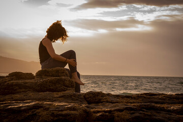 Fototapeta na wymiar young girl sitting down comfortly on the rocks in the beach at sunset. Golden hour