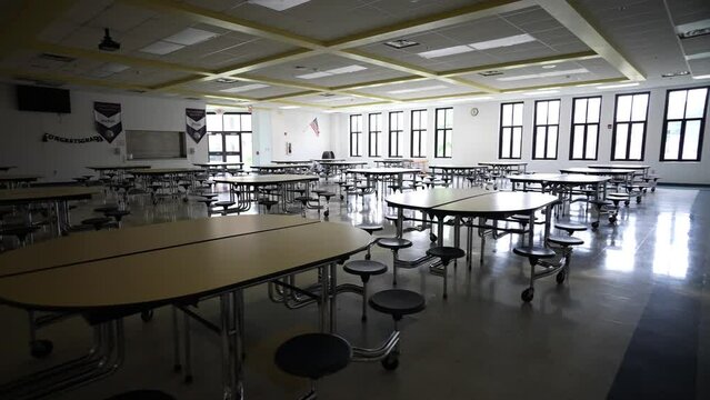 Trucking view to right showing dark and empty school cafeteria with tables and seats.