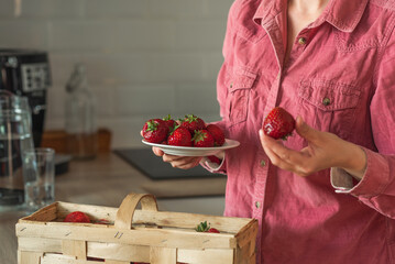 A woman picks strawberries from a basket standing in the kitchen. Strawberries in the hands of a woman.