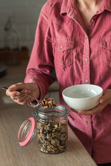 A mature woman puts homemade granola or muesli from a jar in a bowl. The concept of a healthy...