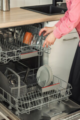 Close-up of female hand loading wished to, empty out or unloading from open automatic built-in dishwashers machine with clean utensils inside in modern home kitchen.