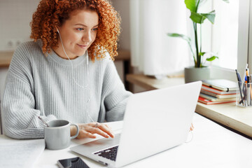 Portrait of curly haired redhead caucasian female in wired headphones spending leisure time using laptop during coffee break, sitting at kitchen table with smartphone, visiting popular web pages - 619194622