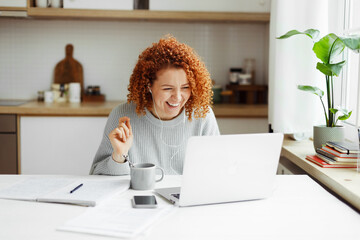 Funny laughing redhead female tutor having video chat with her student, sitting at kitchen table in front of laptop with copy book, coffee cup and smartphone on desk, explaining new topic - 619194645