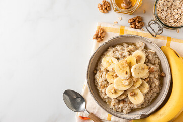Banana, walnuts and honey oatmeal porridge in a bowl with a spoon on white table for healthy tasty...