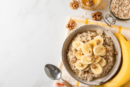 Banana, walnuts and honey oatmeal porridge in a bowl with a spoon on white table for healthy tasty breakfast. Top view