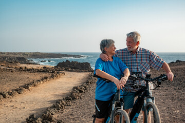 Happy senior couple of cyclists running bicycles in footpath at sea looking each other. Elderly man...