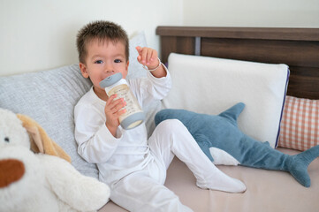 Toddler having his morning milk while sitting on the bed drinking from a bottle with a straw...