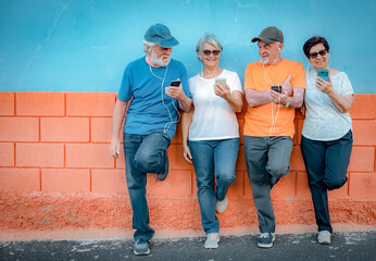 Cheerful group of senior people leaning against an orange and blue wall wearing earphones looking...