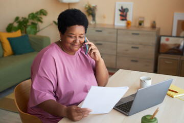 Smiling aged businesswoman with financial document talking to her employer on mobile phone while...